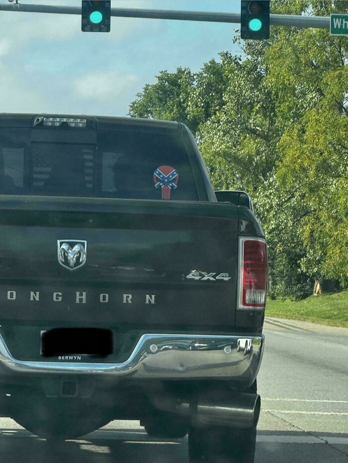 A rear view of a black pickup truck with a cringey confederate flag skull decal on the back window.