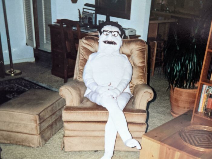 A truly bizarre homemade doll with glasses and a mustache sits cross-legged on a plush armchair in a living room.