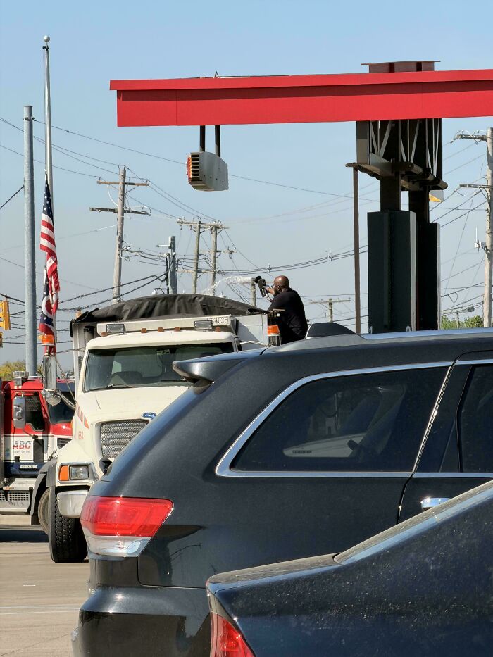 A truly bizarre scene of a person on a white truck bed holding a hose, spraying something next to cars and power lines.