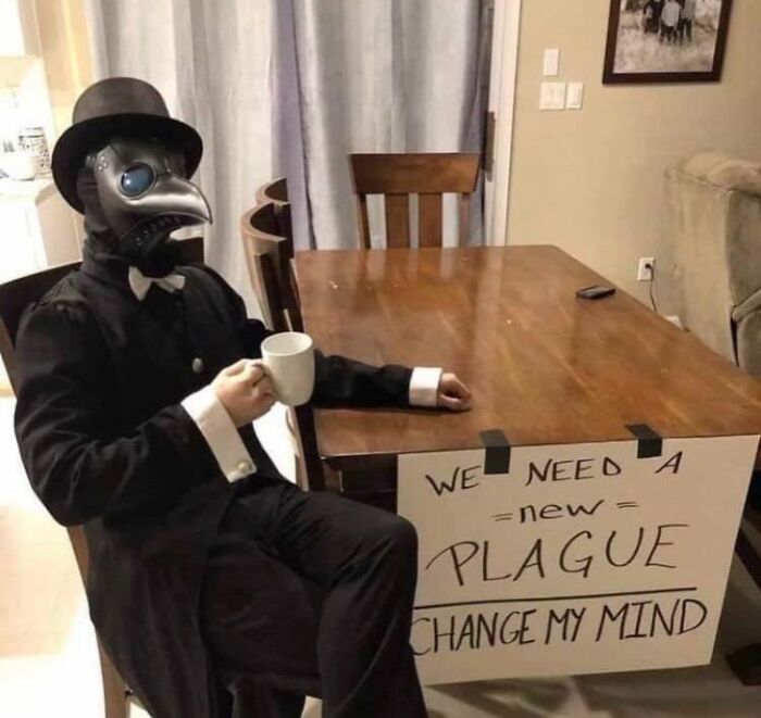 Plague doctor costume sitting at a table with a sign saying We Need A New Plague, demonstrating beliefs and words aging.