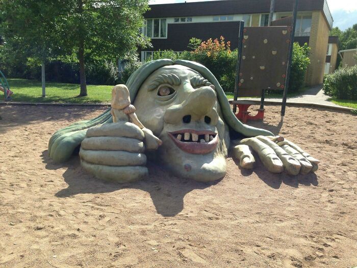 A giant, grotesque troll head statue emerging from sand with a small figure in its hand, depicting playground fails.