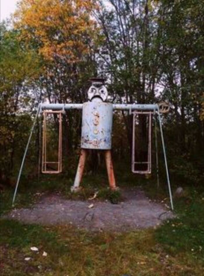 A creepy, old, rusty robot-like structure with swings for arms in a wooded area, highlighting playground fails.