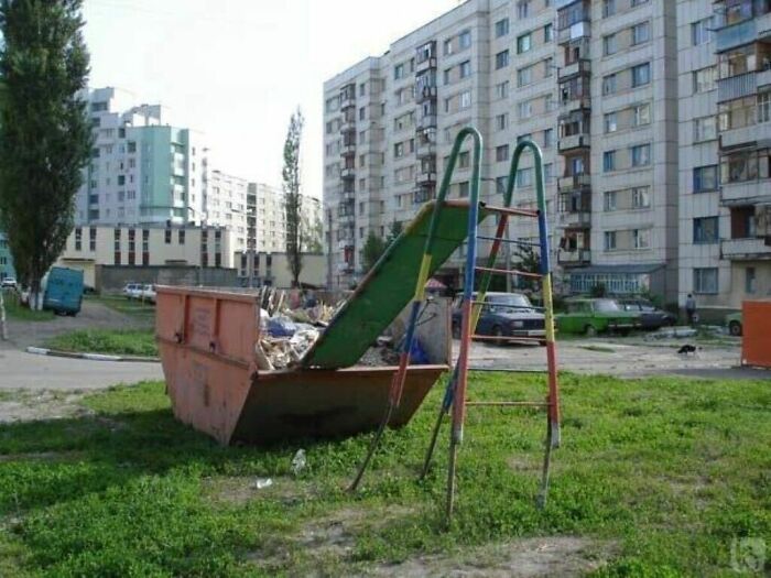 A green playground slide emptying into a large dumpster filled with trash, a clear playground fail.