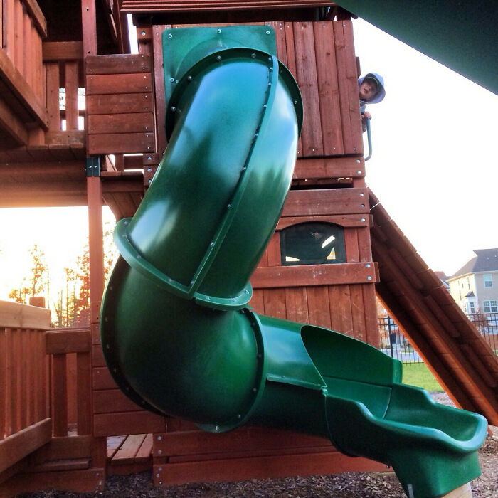 A child peering out from a wooden playground structure with a green, winding slide, highlighting a potential playground fail.