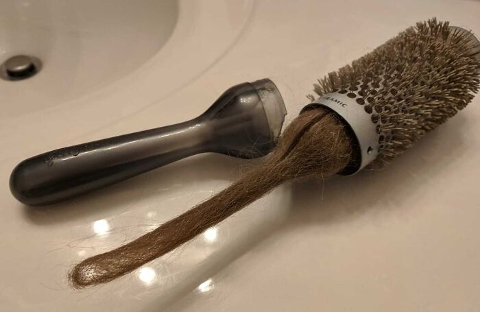 A truly bizarre clump of hair wrapped around a round hairbrush, resembling a rat tail, on a white sink.