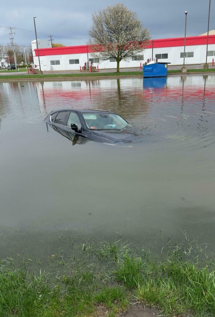 A truly bizarre scene of a black car submerged halfway in a large puddle or flooded area, almost like a lake.