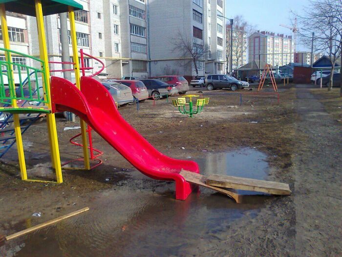 A red playground slide ends in a muddy puddle with a wooden plank. This playground fail looks designed by someone who hates children.