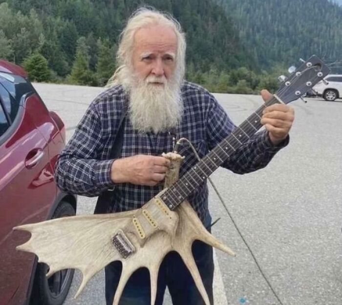 An old man with a long white beard playing a truly bizarre guitar made partly from a large moose antler.