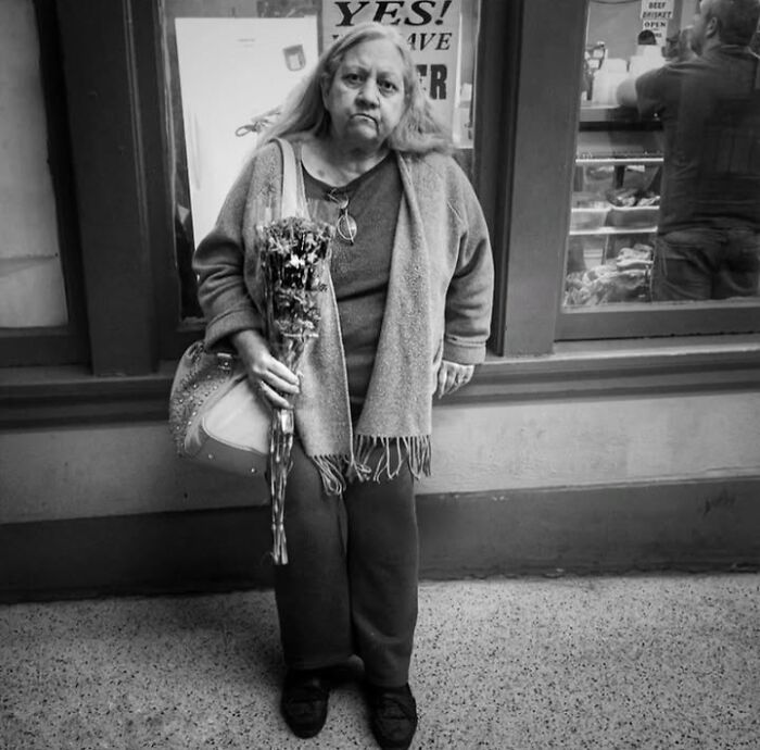 A woman with a bouquet stands against a shop window in a black and white street photo by Taylor Coble.