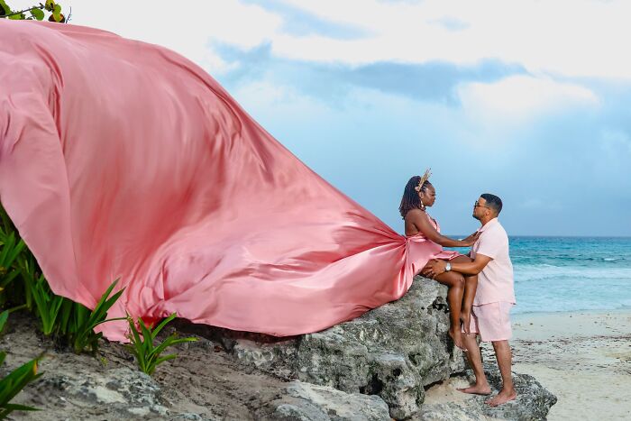 A couple sits on a rock by the ocean, the woman in a flowing pink dress. Proof of finding the right man.