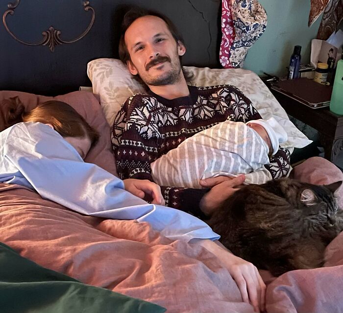 A man smiles holding a swaddled baby in bed with a woman and cat, sweet proof of finding the right man.