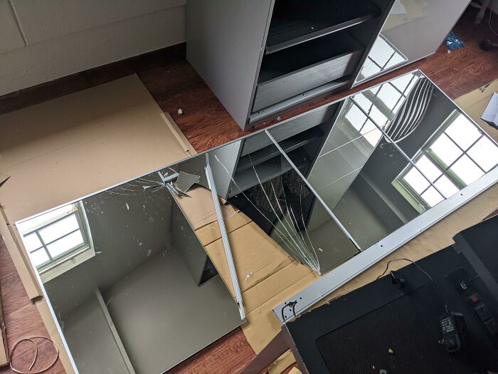 Broken mirrors from a cabinet lie on cardboard, reflecting a window, showing questionable home improvement results.