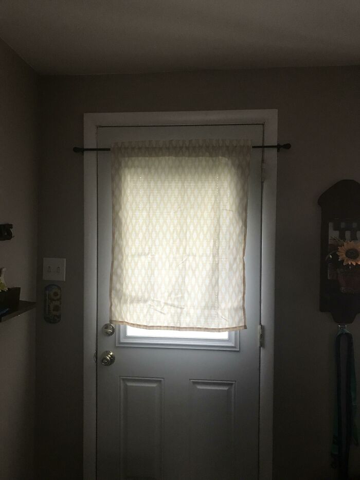 Questionable home improvement project: a small, ill-fitting curtain hung on a door window with a dark room interior.