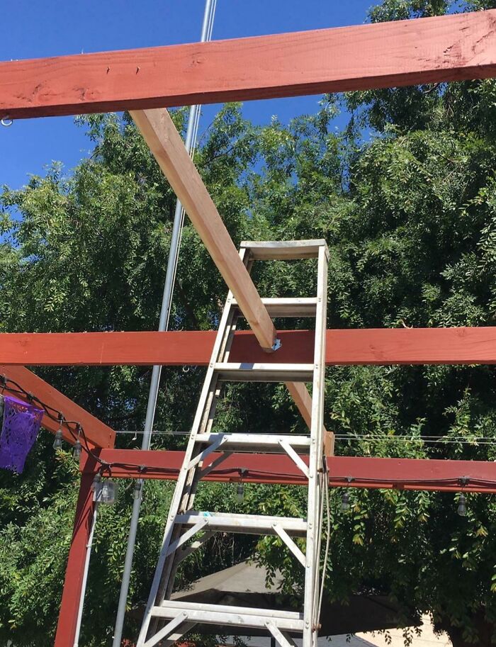 A questionable home improvement project with a wooden beam precariously balanced on a ladder, supporting a new red pergola frame.