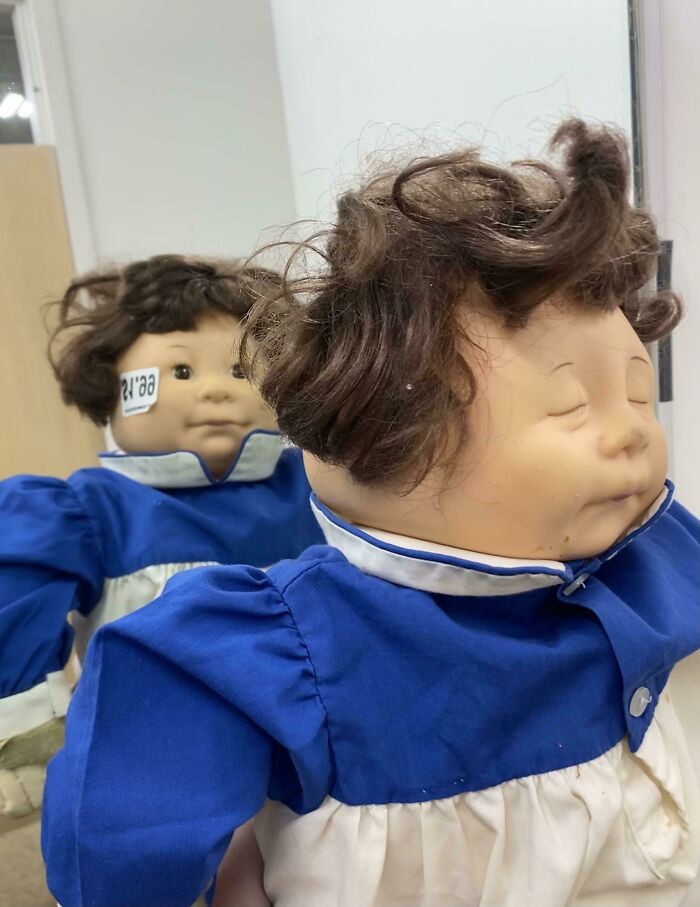 A doll with messy brown hair and a blue dress, reflected in a mirror, one of many unhinged secondhand finds.