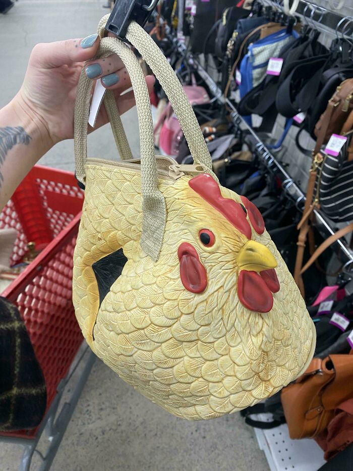 A hand holding a quirky chicken-shaped secondhand purse in a thrift store aisle with other bags in the background.