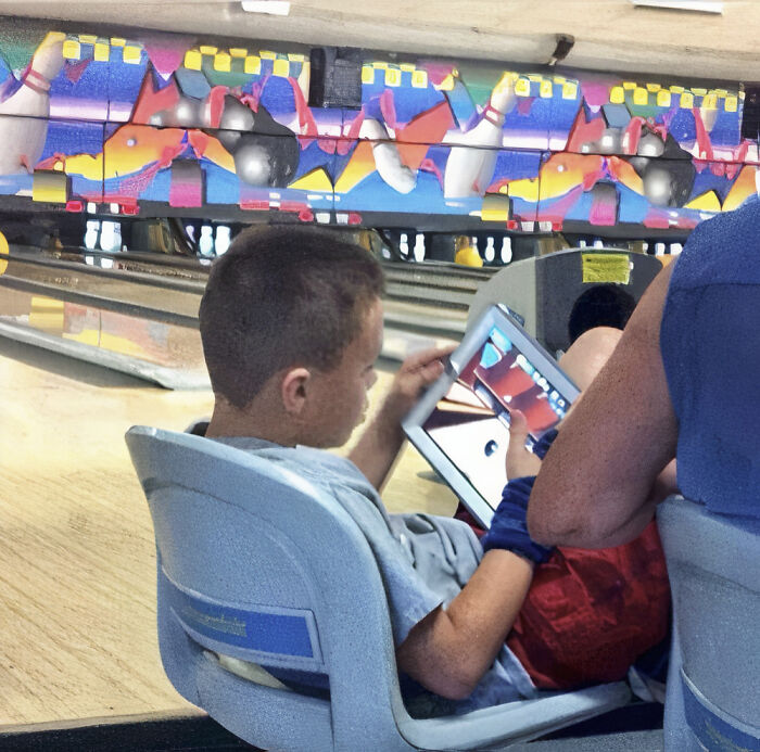 A boy sits in a bowling alley chair playing on a tablet, ignoring the game, a hilarious photo.
