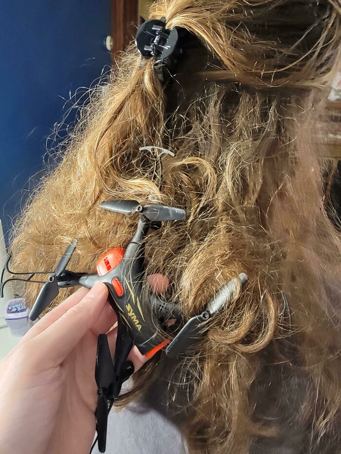 A hand holding a small drone tangled in a child's long, light brown, mischievous hair, highlighting hilarious kids.