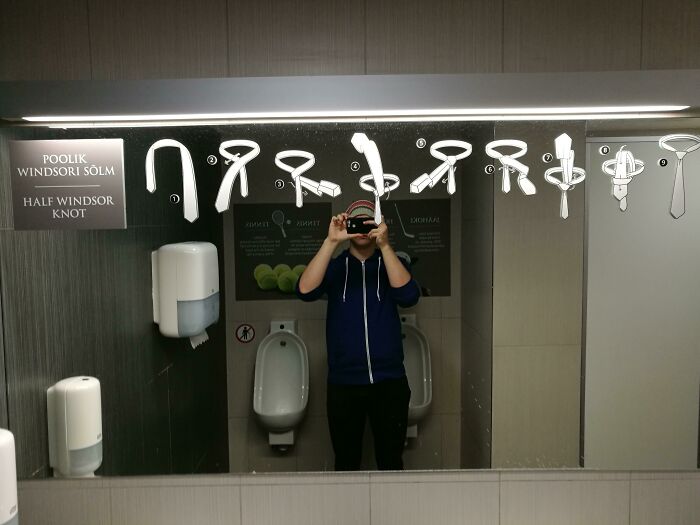 A man takes a selfie in a public restroom mirror with tie-tying instructions, an example of practical product designs.