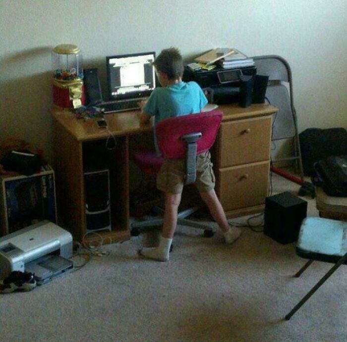 A young boy awkwardly sitting at a computer desk with a printer nearby, an unhinged image that makes people go Hmmm.