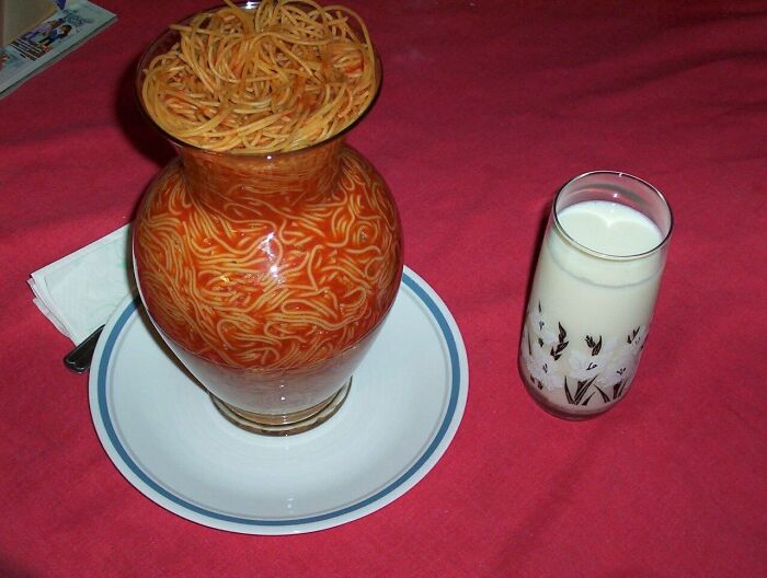 Unhinged images: A glass vase filled with spaghetti and sauce, served on a plate next to a glass of milk.