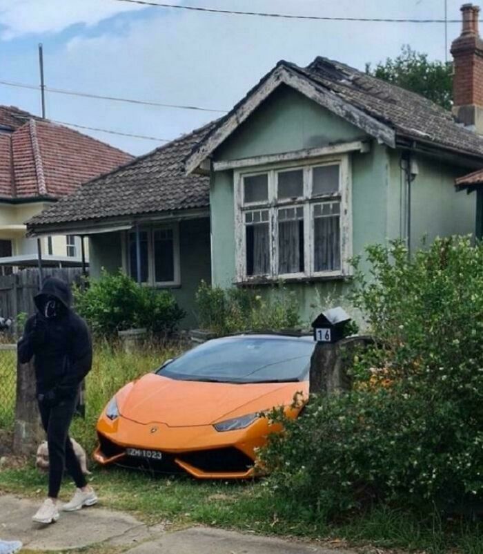 An orange Lamborghini parked by a run-down house, creating an unhinged image.