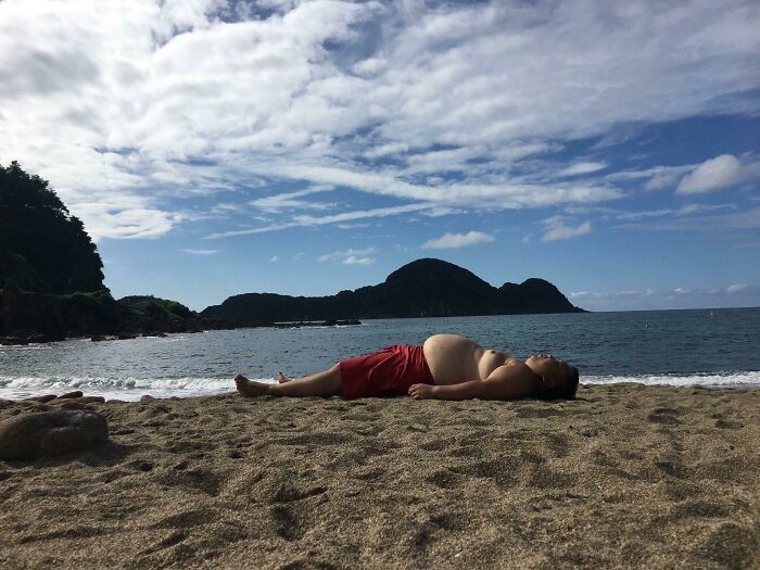 A man with a large belly lays on his back on a sandy beach next to the ocean under a cloudy sky, showcasing unhinged images.