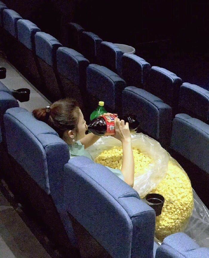 A woman in a movie theater seat drinks from a large soda bottle next to a huge bag of popcorn, one of many unhinged images.
