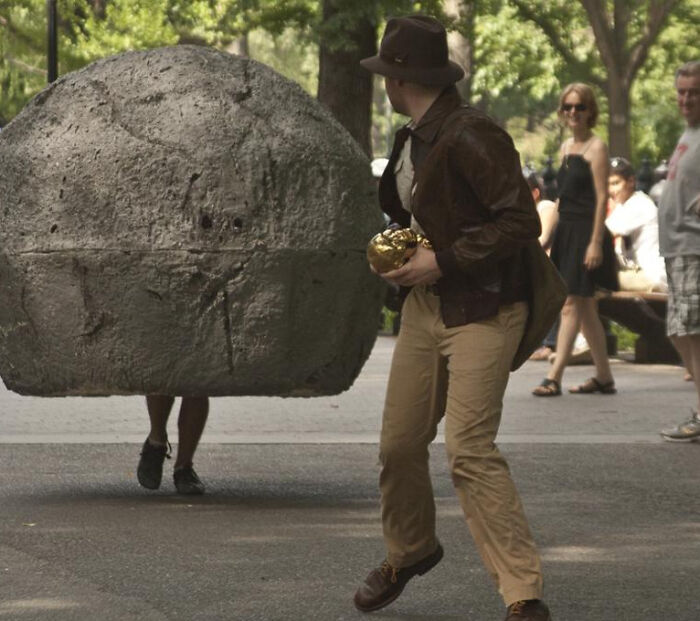 A person dressed as Indiana Jones running from a large rolling boulder prop, an unhinged image.