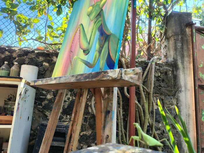 A vibrant painting of a praying mantis on an easel outdoors, amazingly synced just right with a real mantis on the ground.