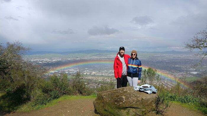 A couple stands on a hill with a city and a rainbow perfectly synced in the background.