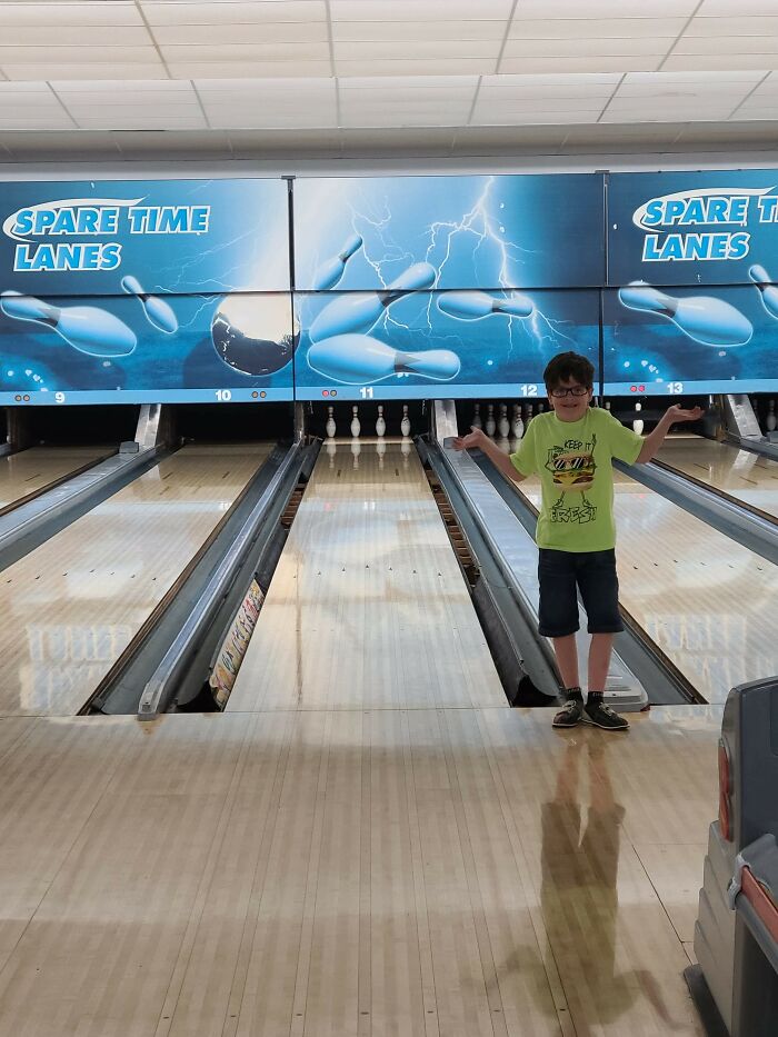 A boy standing on a bowling alley lane, arms out in a shrug, with bowling pins in the background, a moment synced just right.
