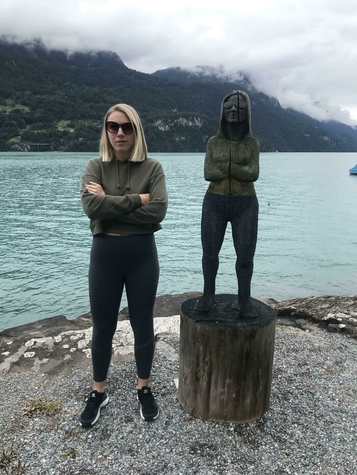 A woman standing next to a statue that perfectly matches her pose by a lake, a moment synced just right.