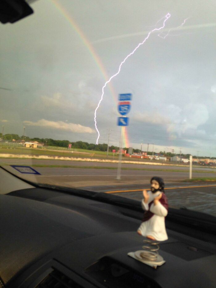 A bobblehead Jesus on a car dashboard, with a rainbow and lightning strike outside, a moment synced just right.