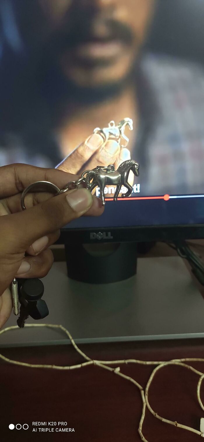 A hand holding a horse keychain, perfectly synced with a horse shown on a television screen in the background.