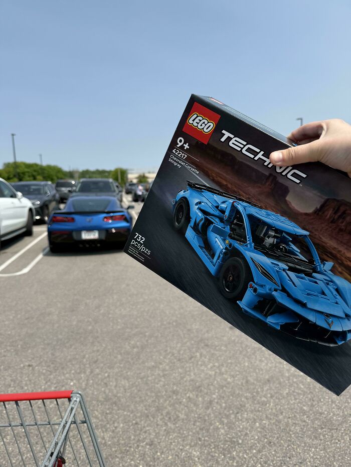 A person holding a LEGO Technic Chevrolet Corvette box, perfectly aligned with a real blue Corvette in the background, something synced just right.