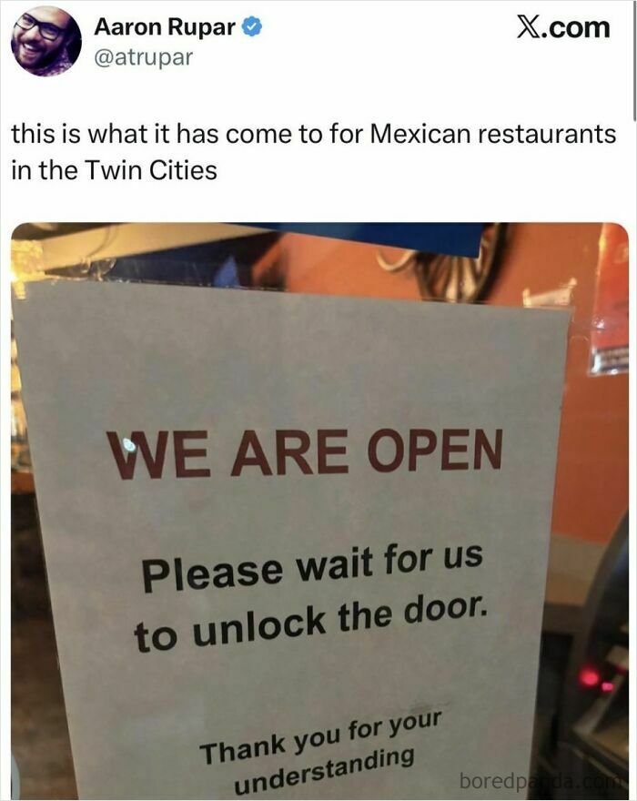 A sign in a restaurant window stating 'WE ARE OPEN Please wait for us to unlock the door,' portraying a dystopian nightmare of business challenges.