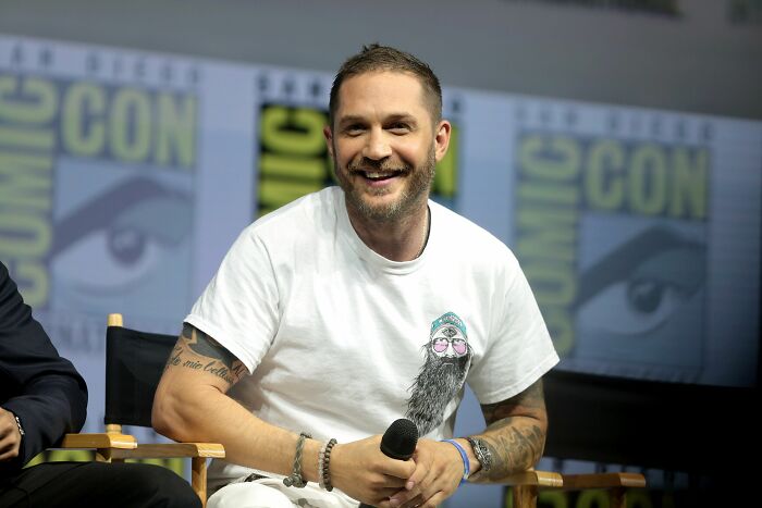 Tom Hardy smiling, wearing a white t-shirt, holding a microphone, with a Comic-Con banner in the background. An actor's career highlight.