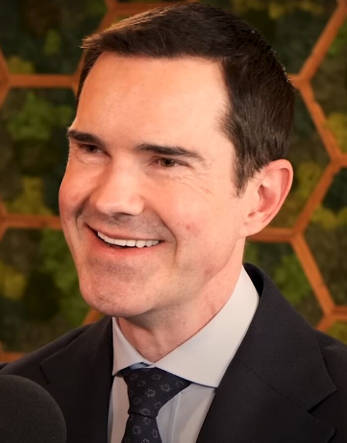 A smiling Jimmy Carr, known for his career highlights in comedy, wearing a suit and tie against a green background.