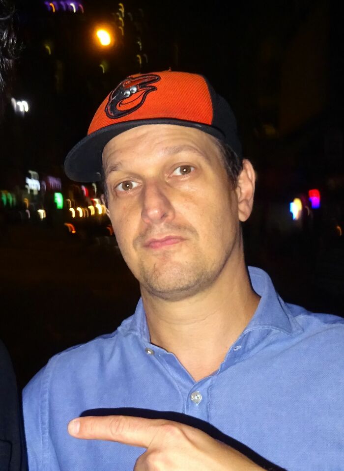 A close-up of Josh Charles, a man in a blue shirt and an Orioles baseball cap, pointing with his right hand.