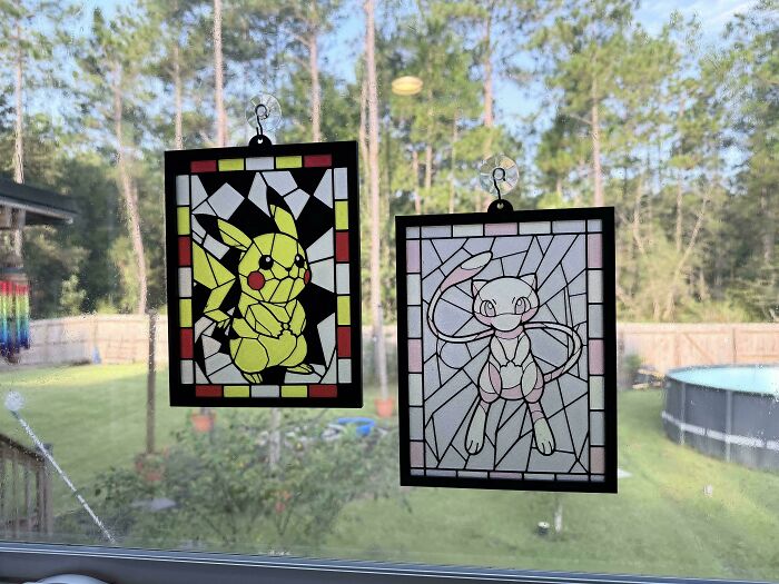 Two framed 3D printed stained-glass style images of Pikachu and Mew hanging on a window.