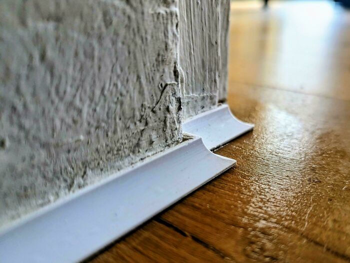 A close-up of a white baseboard expertly fitted against a textured wall and wooden floor, an item people 3D printed.