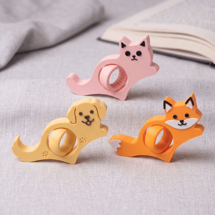 Colorful animal-shaped book page holders, including a pink cat, yellow dog, and orange fox, all printed with 3D printers.