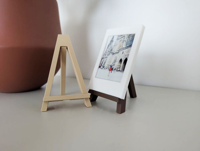 Two small, 3D printed easels, one holding a miniature framed photo, on a light grey surface.