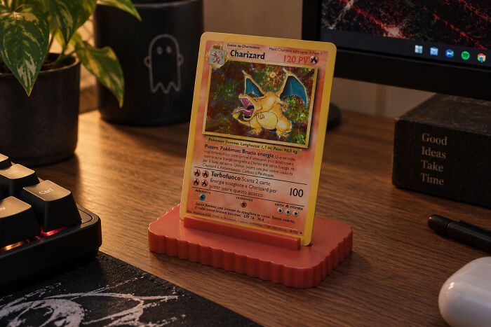 A 3D printed red stand holding a Pokemon Charizard card on a wooden desk with a plant and keyboard nearby.
