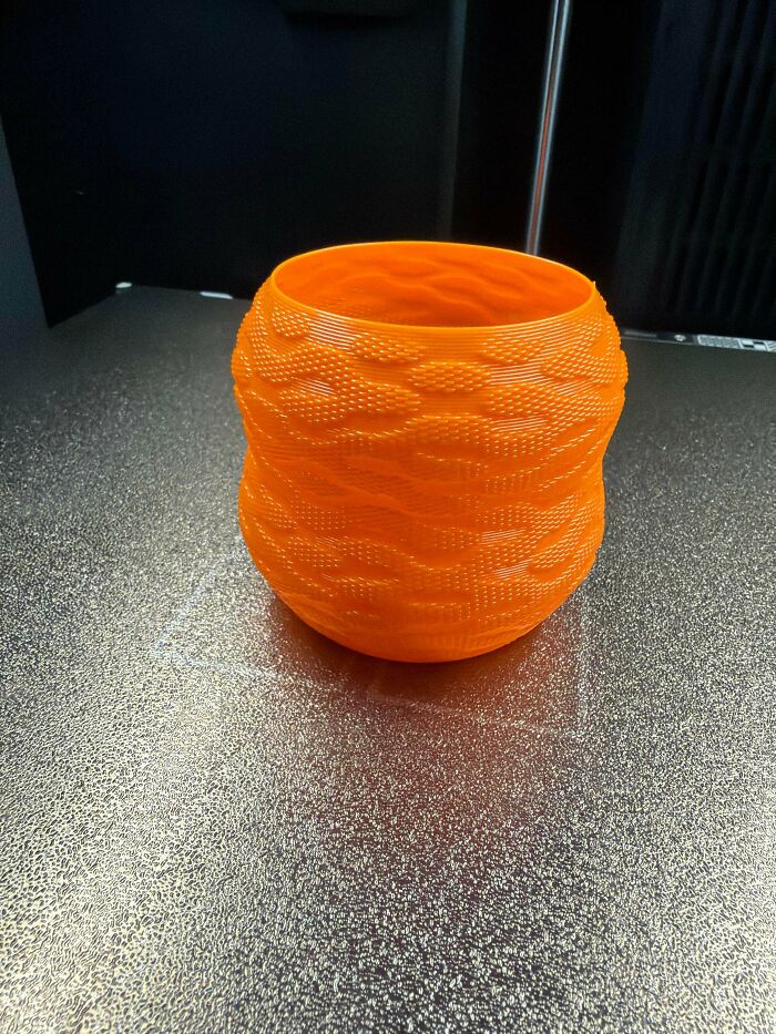An orange, textured vase with a wavy pattern, created with 3D printers, sitting on a shiny surface.
