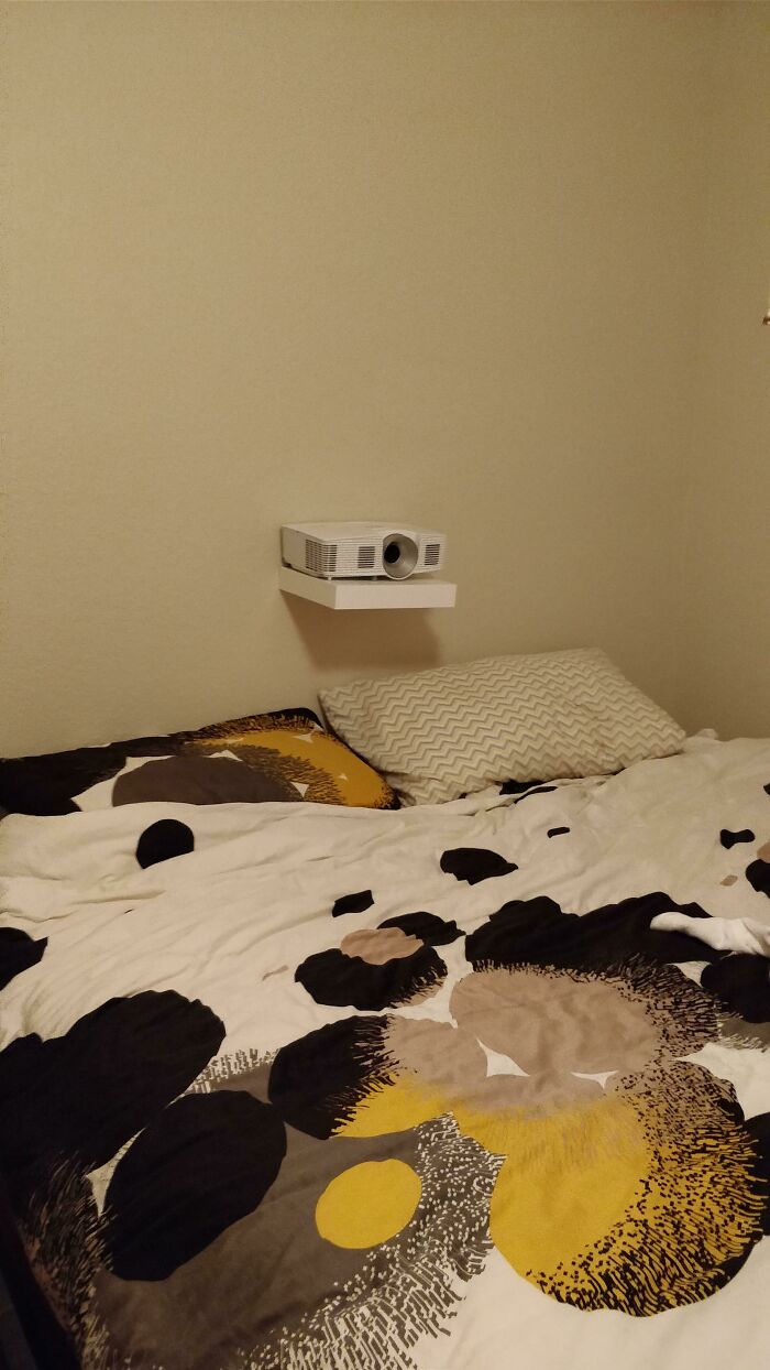 A DIY fail of a projector precariously placed on a small, wall-mounted shelf above a bed.
