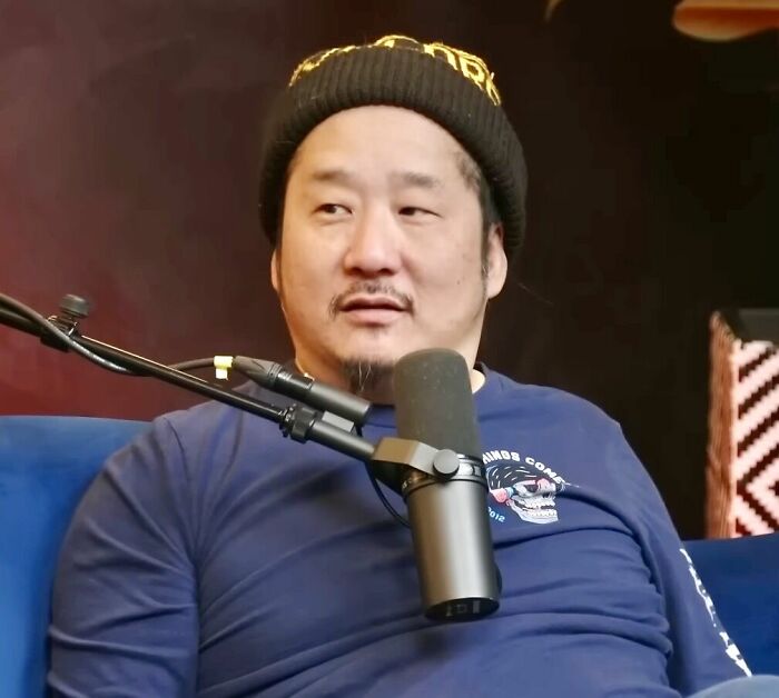 Comedian Bobby Lee, wearing a black beanie and blue long-sleeved shirt, sitting in front of a microphone, looking to the side.