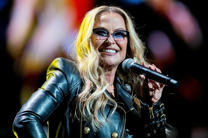 Anastacia smiling, wearing sunglasses and a black leather jacket, holding a microphone during a career highlight performance.