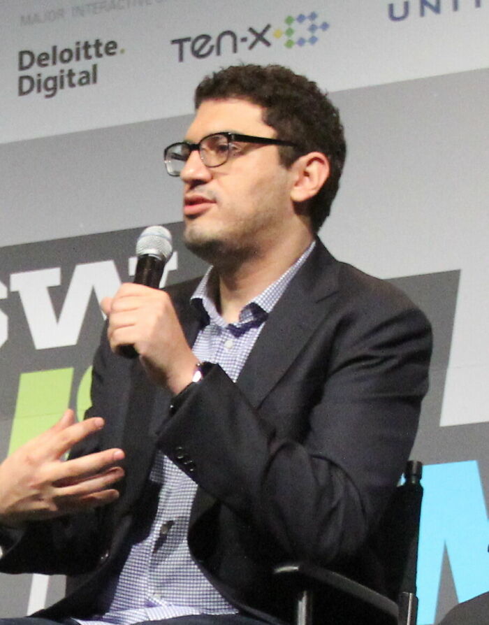 Sam Esmail, a director, wearing glasses and a suit, speaks into a microphone at an event, highlighting his career.